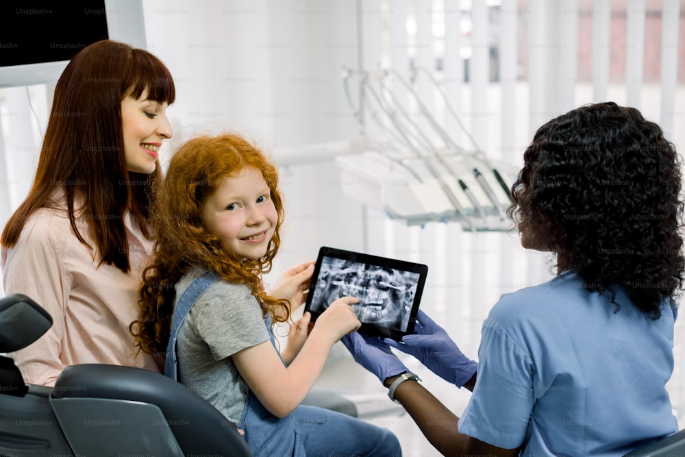 Back view of mother and child girl at appointment with female black doctor orthodontist, showing xray teeth scan on tablet. Dental examination and dentist consultation. Little girl looks at camera.