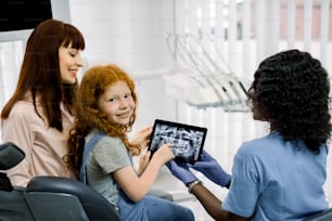 Vista posterior de la madre y la niña en la cita con la ortodoncista doctora negra, que muestra el escaneo de los dientes de rayos X en la tableta. Examen odontológico y consulta odontológica. La niña mira a la cámara.