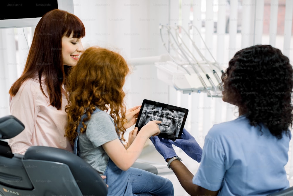 Young African woman dentist showing dental x-ray on tablet to his little red haired girl patient and her mother before medical procedure in clinics. Pediatric dentistry and orthodontics.
