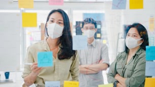 Jovens empresários da Ásia discutindo reuniões de brainstorming de negócios trabalhando juntos compartilhando dados e escrevendo parede de vidro com máscara facial médica de volta ao trabalho no escritório. Vida e obra após o coronavírus.