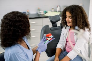 아프리카 여성 의사가 어린 아이 환자인 혼혈 여학생에게 적절한 치과 치료를 시연하고 있습니다. 현대 치과 의사의 사무실에서 듣고 즐거운 시간을 보내는 행복한 아이.