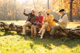 Senior people using mobile smart phones in beautiful autumn park. One elderly man reading a book. Retired friends spending time together outdoor.