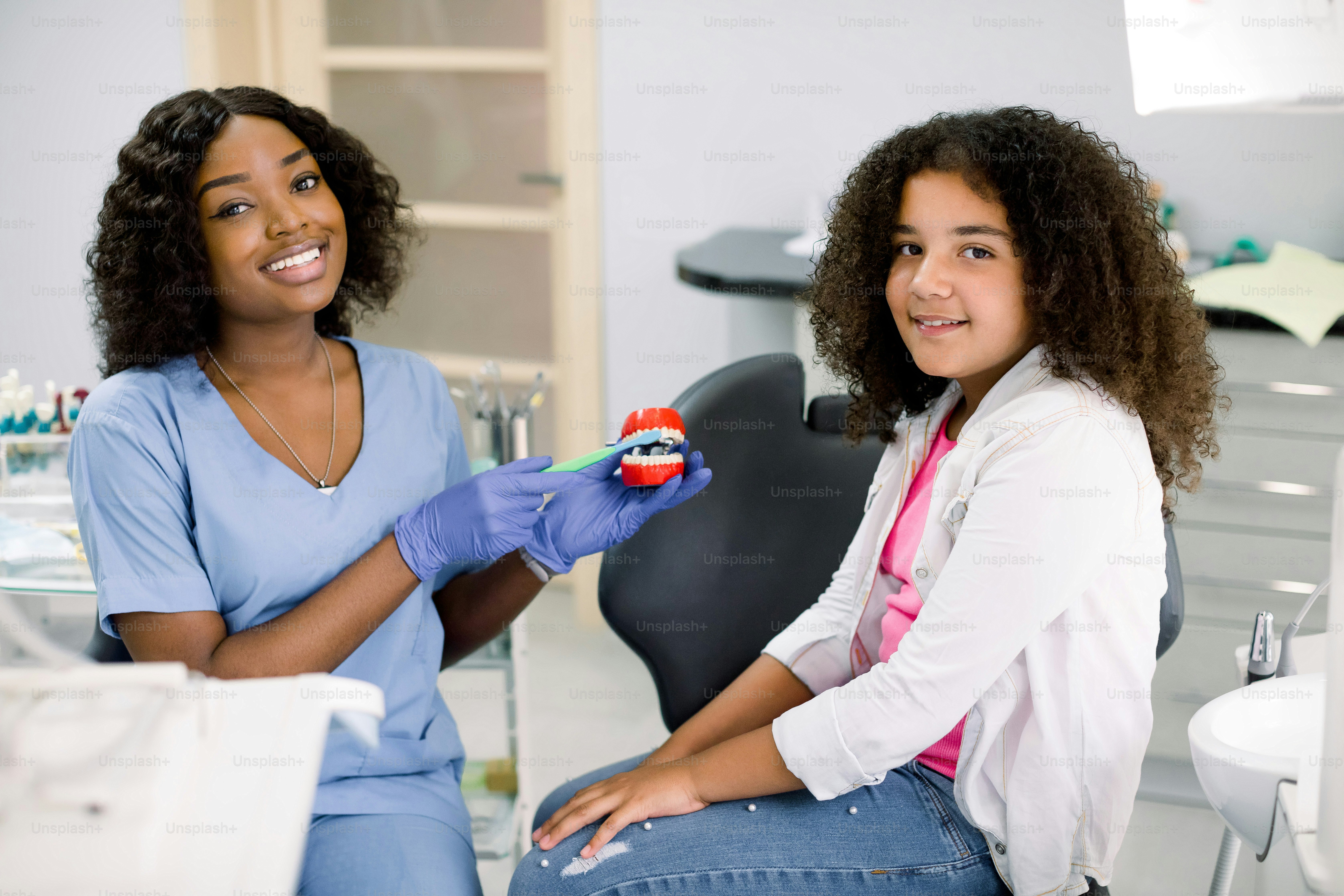 Happy smiling girl at dentist's office. Cute mixed raced a curly girl, sits in dental chair and looks at camera, while her female African dentist shows how to properly brush teeth on jaw model.