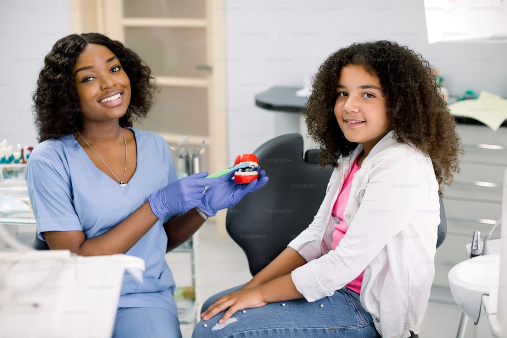Happy smiling girl at dentist's office. Cute mixed raced a curly girl, sits in dental chair and looks at camera, while her female African dentist shows how to properly brush teeth on jaw model.