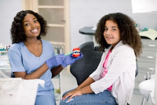 Chica sonriente feliz en el consultorio del dentista. Linda raza mixta una niña rizada, se sienta en el sillón dental y mira a la cámara, mientras que su dentista africana muestra cómo cepillarse correctamente los dientes en un modelo de mandíbula.