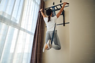 Back view of a sportswoman hanging from the horizontal bar supported with an elastic rope