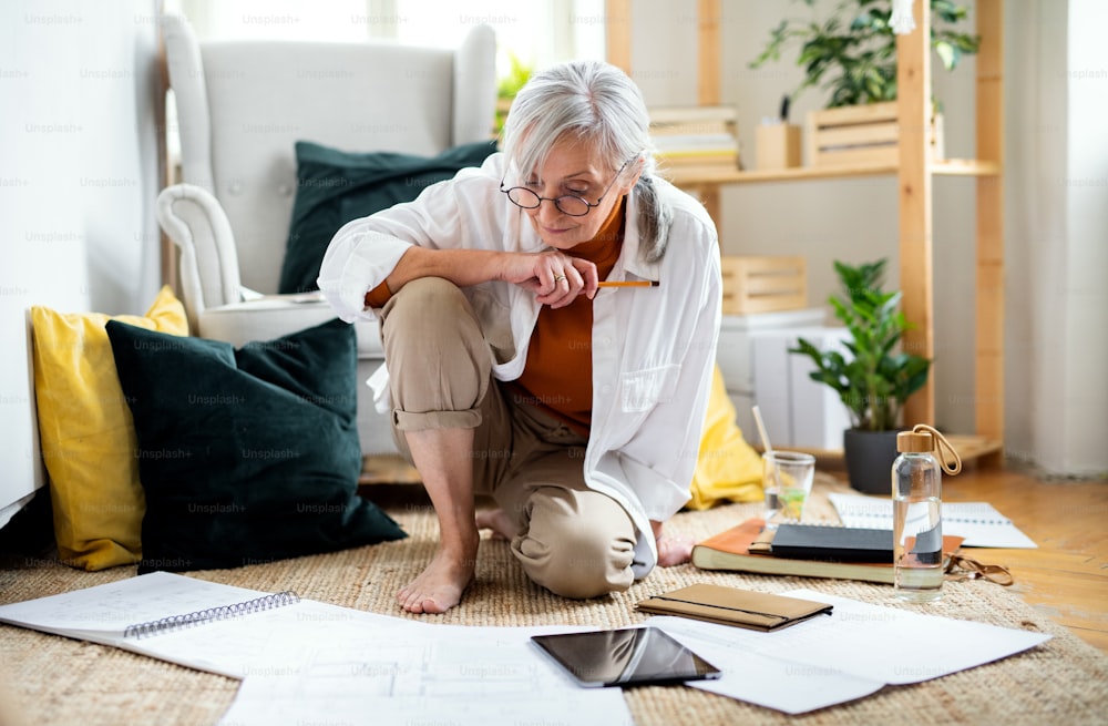 Portrait of senior woman architect sitting with blueprints on floor indoors at home, working.