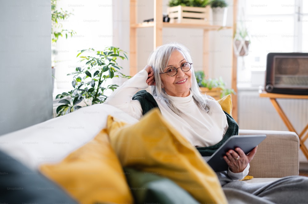 Ritratto di donna anziana seduta all'interno sul divano di casa, rilassante e utilizzando il tablet.