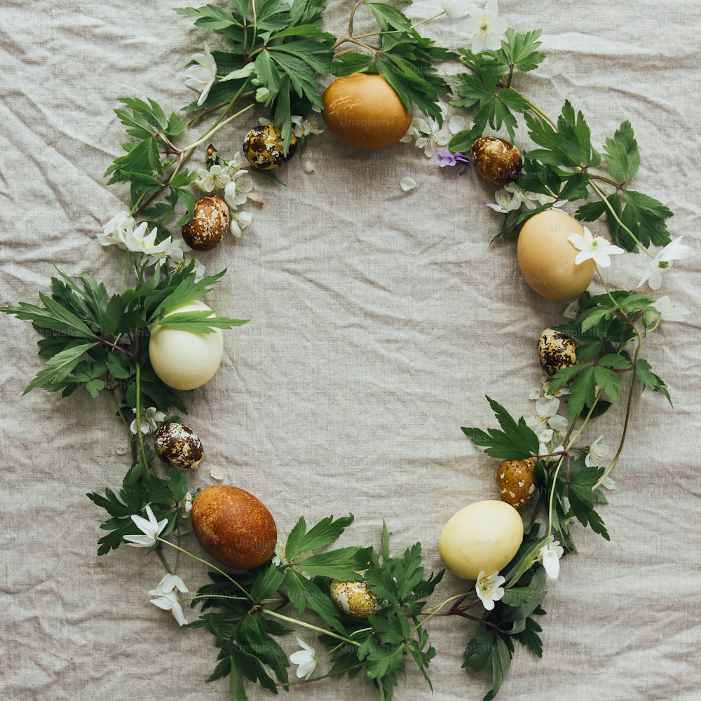 Happy Easter! Easter eggs and spring flowers in wreath on rustic linen, flat lay with space for text. Aesthetic stylish easter eggs in natural dye and blooms, seasons greetings