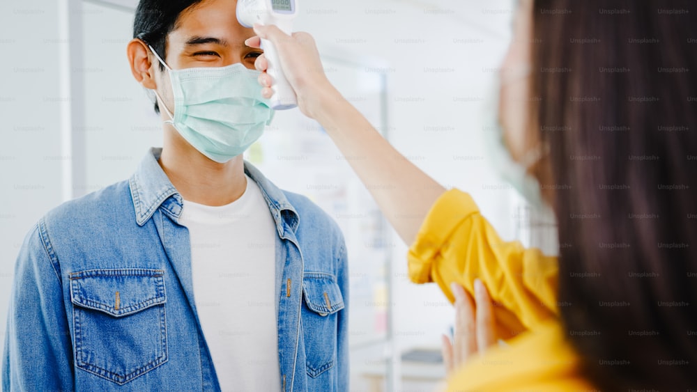 アジアの女性受付係は、オフィスに入る前に、顧客の額に赤外線温度計チェッカーまたは温度ガンを使用して保護フェイスマスクを着用します。コロナ禍後のライフスタイルのニューノーマル。