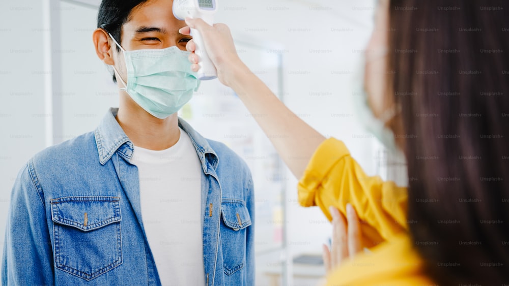 Asia female receptionist conducting wear protective face mask use infrared thermometer checker or temperature gun on customer's forehead before enter office. Lifestyle new normal after corona virus.