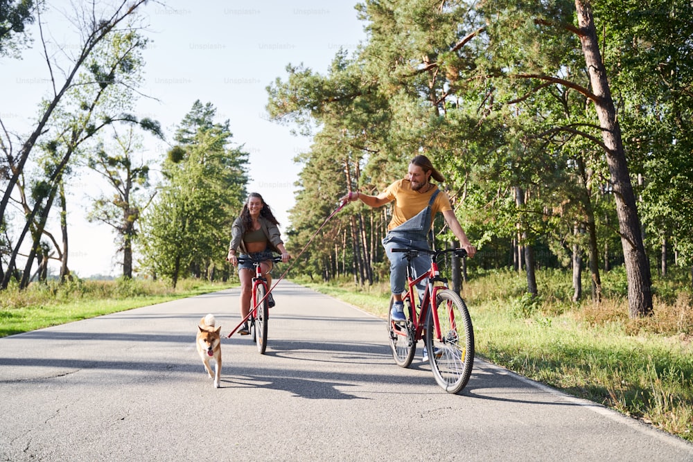 Young man and woman are riding the bike in the park with many trees. Young man holding a leash with a dog running next to him