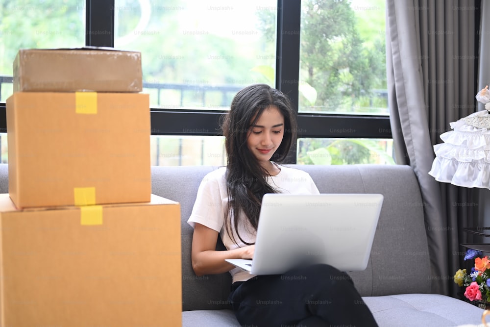 Start up small business entrepreneur SME or freelance woman working with laptop computer sitting surrounded by boxes at home. SME concept.