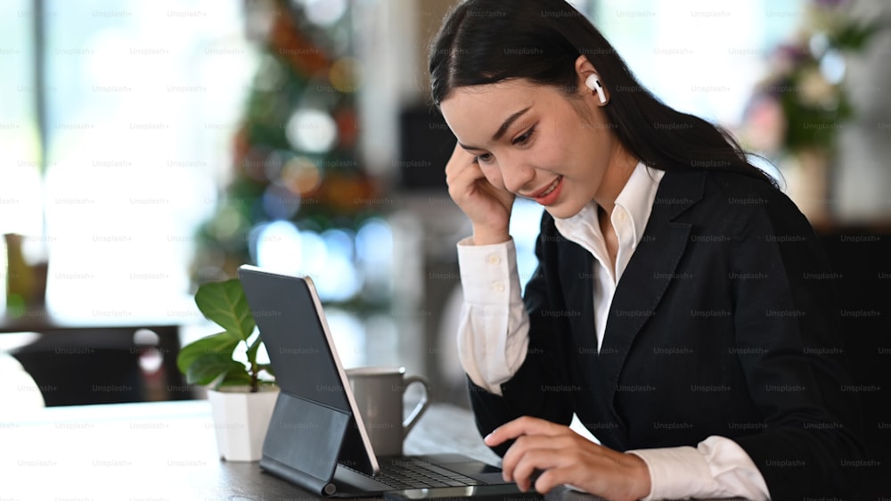 Femme d’affaires en costume formel portant des écouteurs sans fil et travaillant sur une tablette dans un bureau moderne.