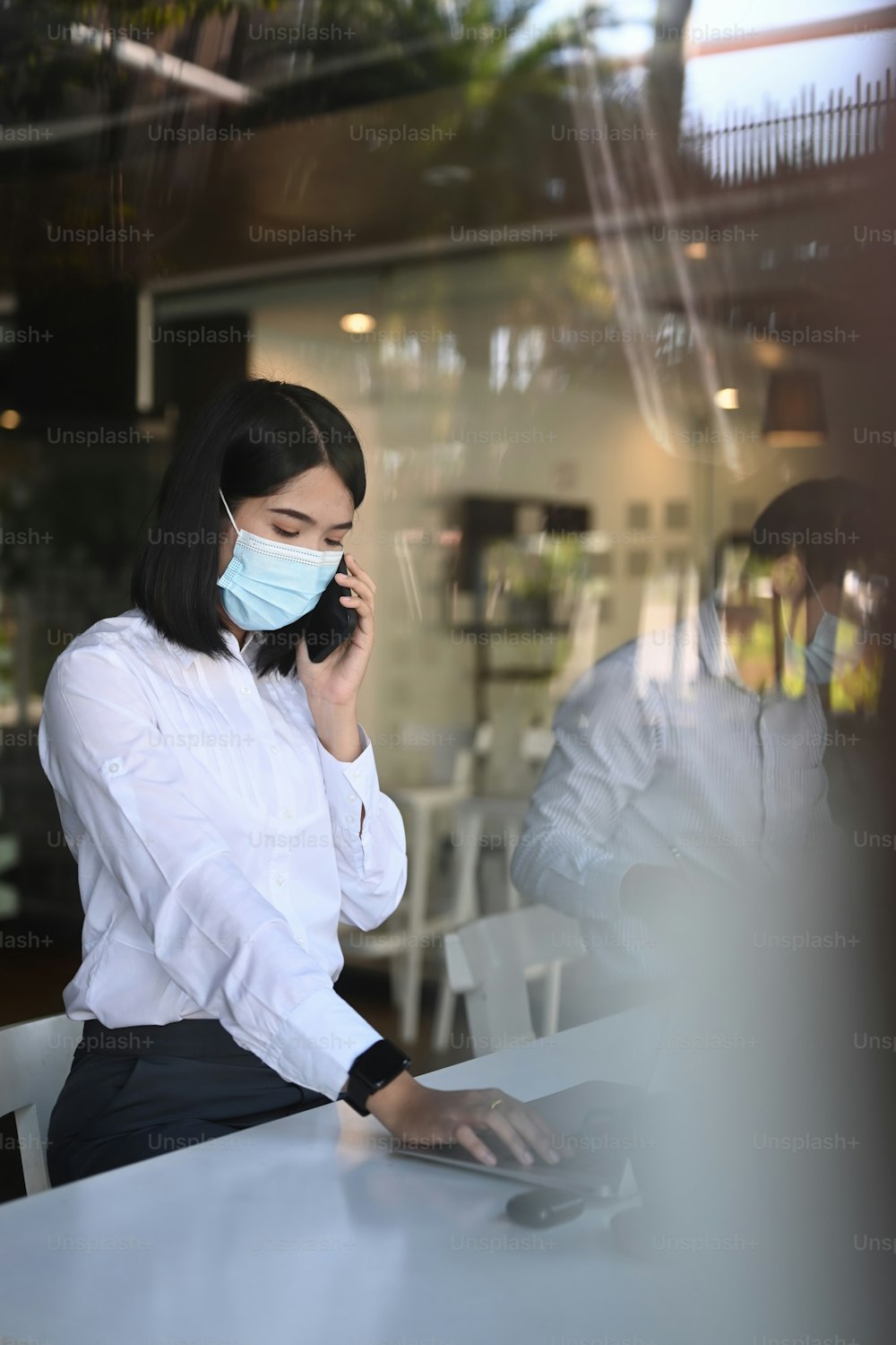 Portrait of businesswoman wearing protective mask working on laptop and talking on mobile phone while her colleague sitting in background.