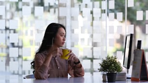 Donna d'affari che distoglie lo sguardo pensierosa e tiene in mano una tazza di caffè mentre è seduta nel suo spazio di lavoro.