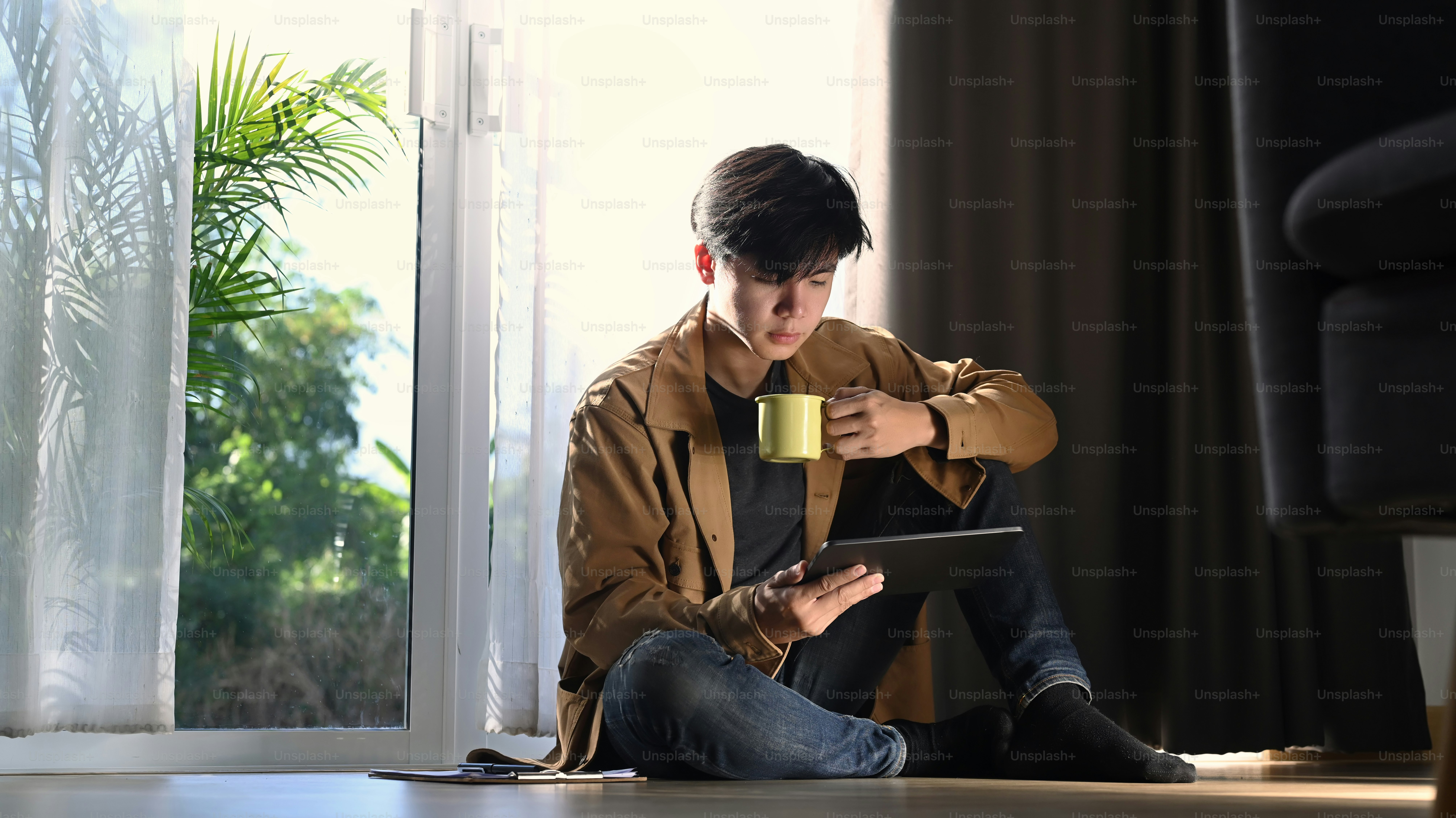 A man sitting on the floor his holding coffee and looking on digital tablet.
