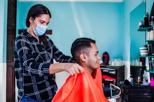 latin woman hairdresser with face mask cutting hair with clipper machine and comb. Mexican man gets a haircut. Barber services with security measures from Covid-19 in Mexico city