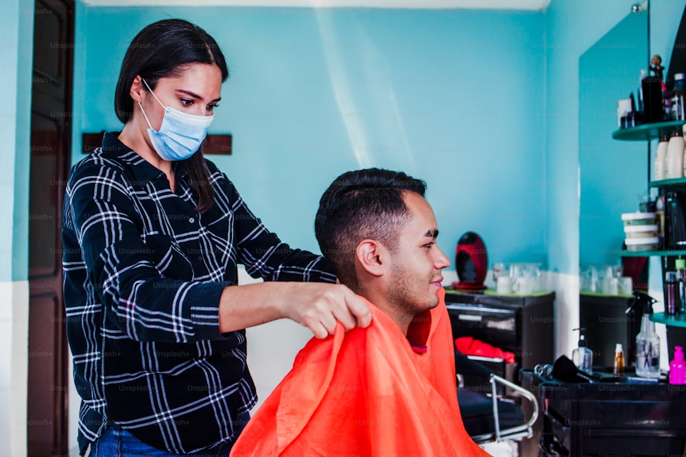 latin woman hairdresser with face mask cutting hair with clipper machine and comb. Mexican man gets a haircut. Barber services with security measures from Covid-19 in Mexico city