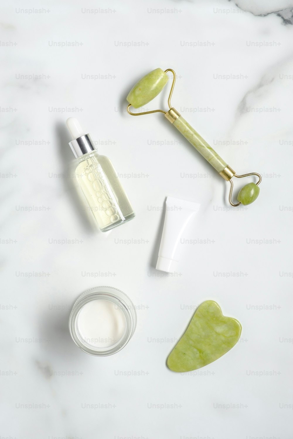 Jade stone facial roller, gua sha, serum lotion, moisturizer, tube with cream on marble desk. Flat lay, top view. Anti-aging cosmetics set.