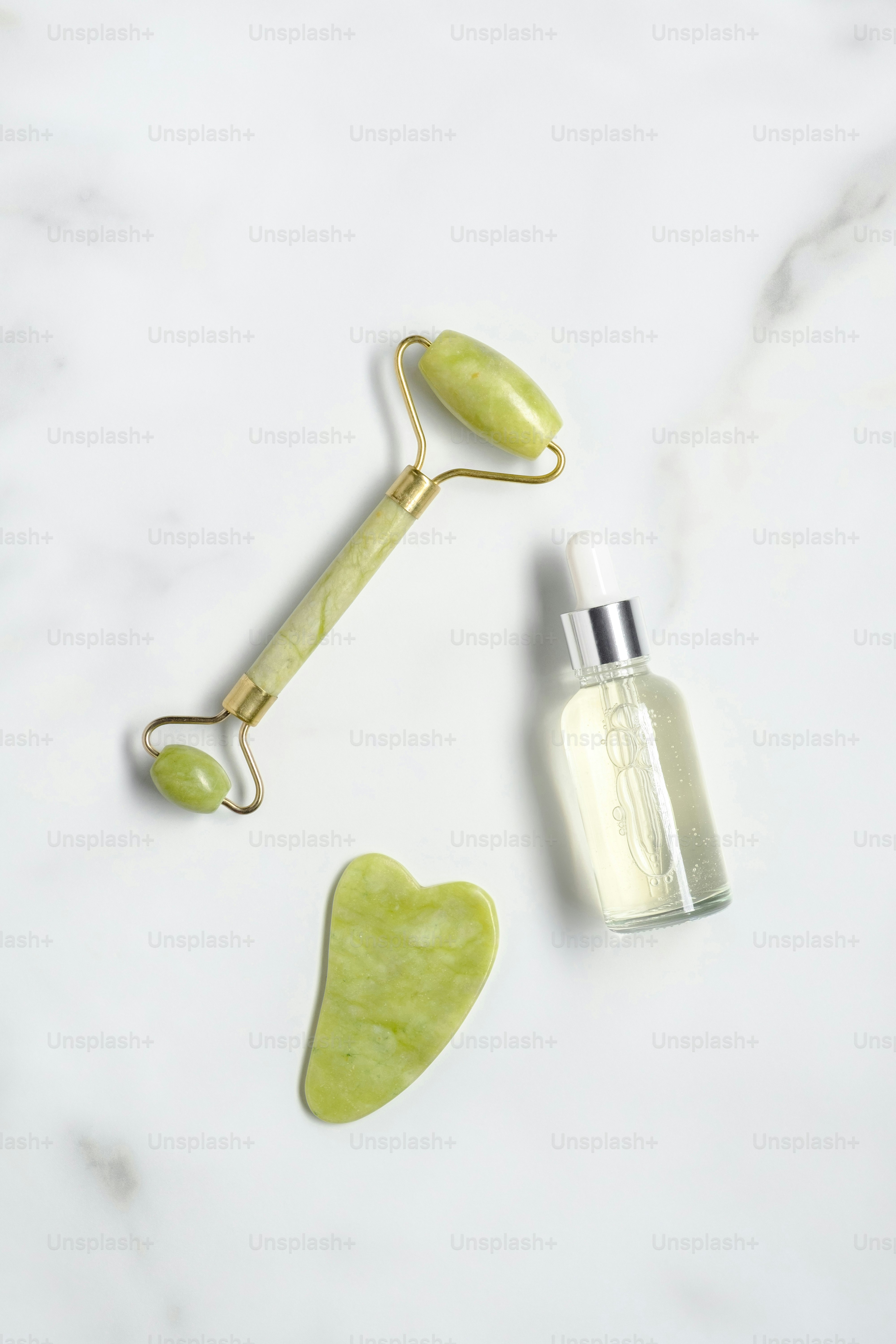 Jade stone facial roller, gua sha, serum lotion on marble table. Facial