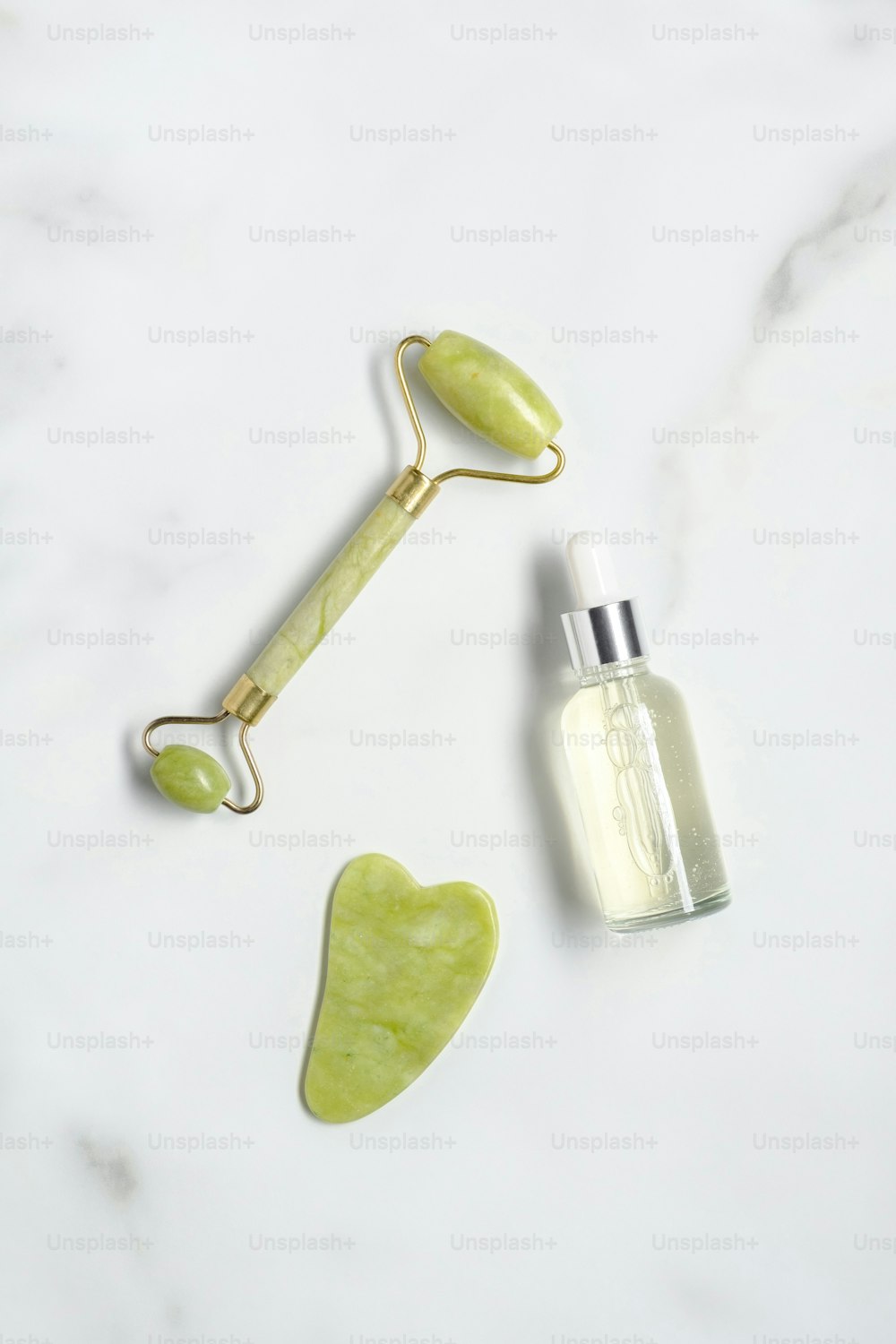 Jade stone facial roller, gua sha, serum lotion on marble table. Facial skin care cosmetics set. Flat lay, top view.