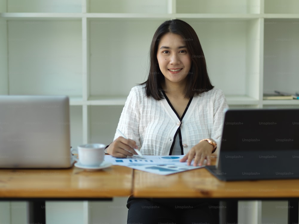 Porträt einer Geschäftsfrau, die beim Lesen von Papierkram im Büroraum in die Kamera lächelt