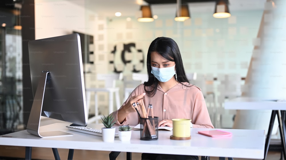 Businesswoman wearing face mask working with computer and analyzing business report in office.