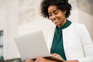 Mujer de negocios afro usando su computadora portátil mientras está sentada en las escaleras al aire libre. Concepto urbano y empresarial.