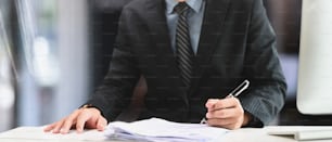 Foto recortada de un hombre de negocios sosteniendo un bolígrafo firmando en el trabajo de papel en el escritorio de la oficina.