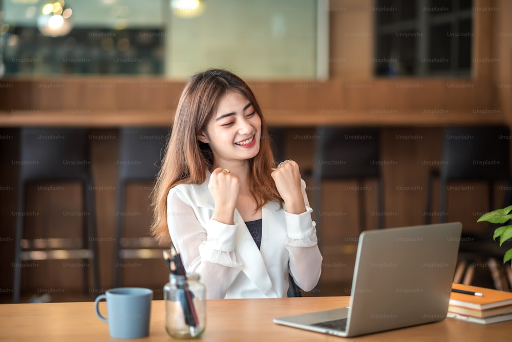 Une femme d’affaires asiatique heureuse célèbre le succès avec le bras levé devant l’ordinateur portable au bureau.