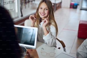 Waist up portrait of funny smiling teenager girl in white shirt choosing and trying different glasses while sitting at the desk in optician center
