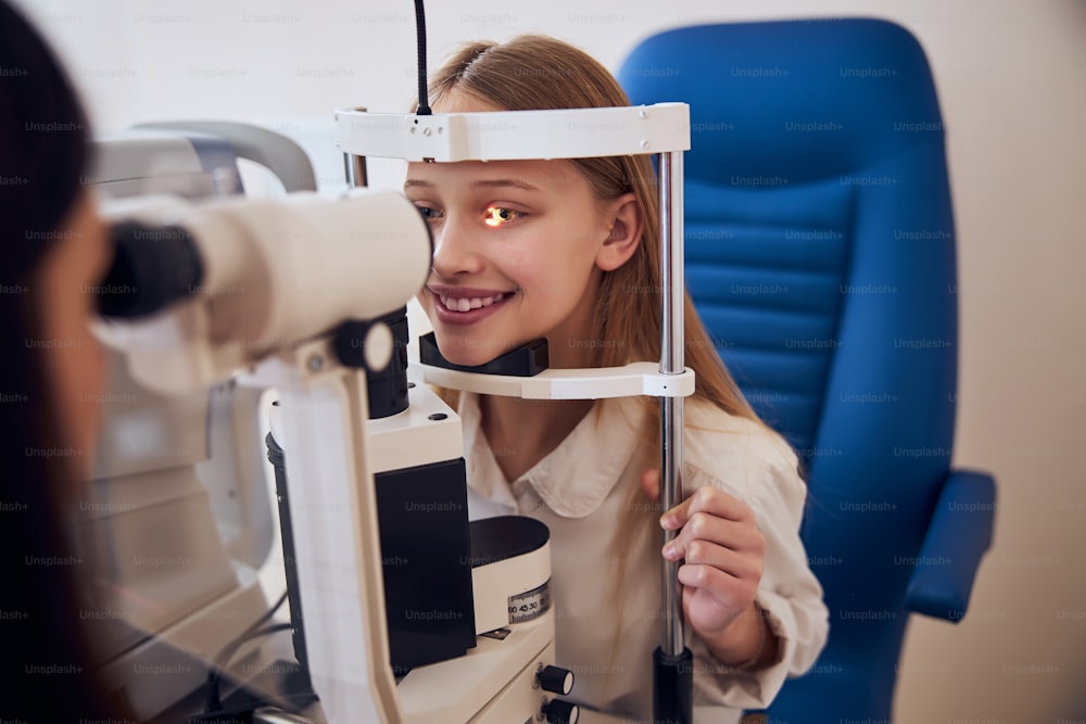 Caucasian female child sitting in blue medicine chair and looking at slit lamp machine during medical check up in eye clime