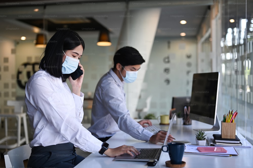 Side view of businesswoman wearing protective mask working on laptop and talking on mobile phone while sitting with her colleague at office.