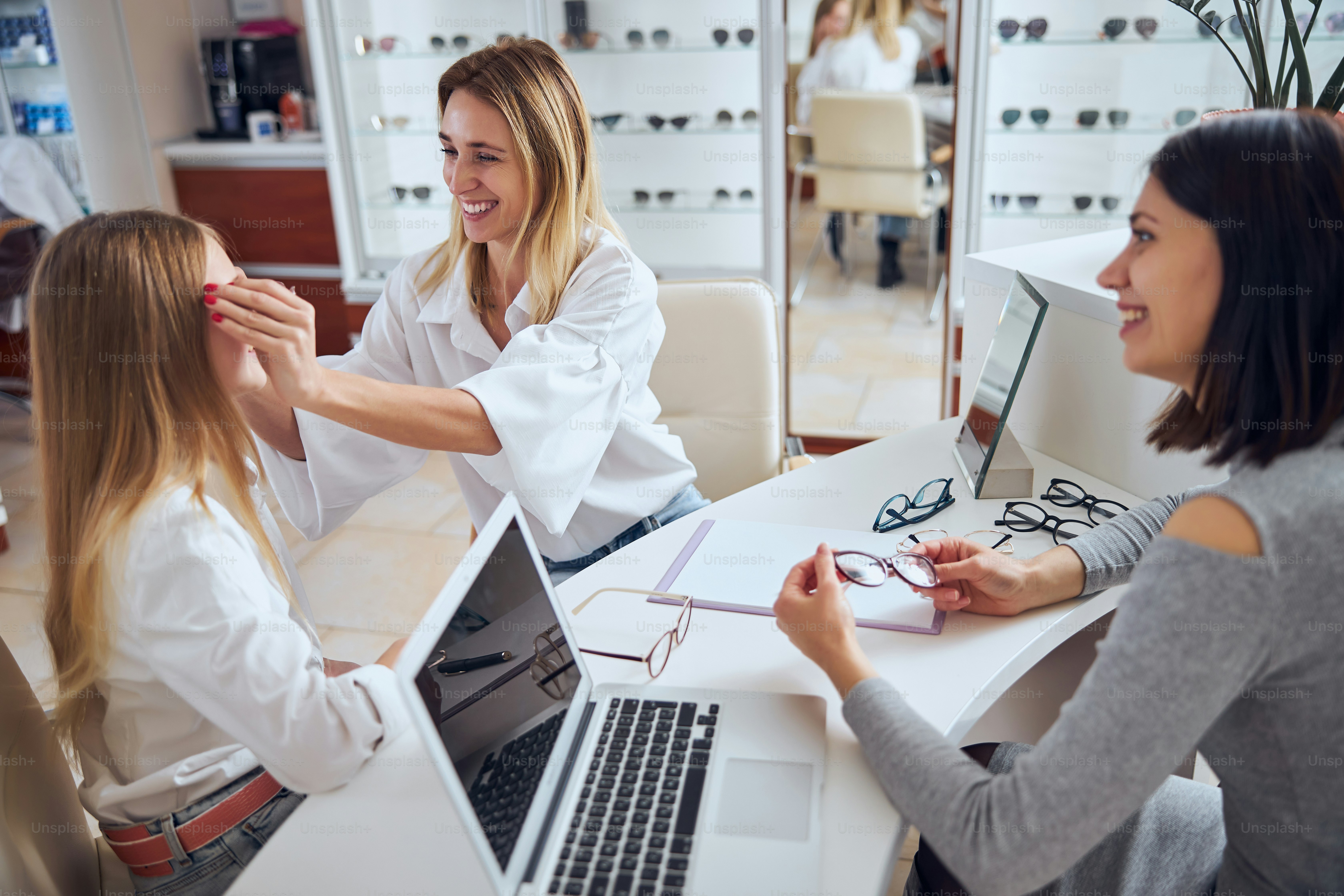 Seitenansicht Porträt des Verkäufers, der verschiedene mehrfarbige Brillen für den Patienten vorschlägt, während die Mutter sie auf dem Gesicht trägt Tochter im Büro Optiker
