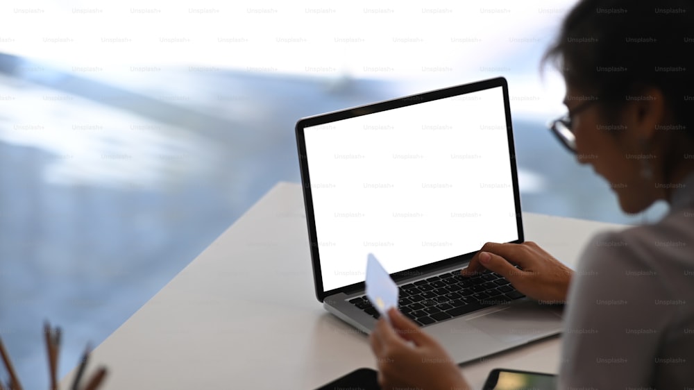 Vista trasera de una mujer joven que usa una computadora portátil y sostiene una tarjeta de crédito que realiza pagos en línea o compras en línea.