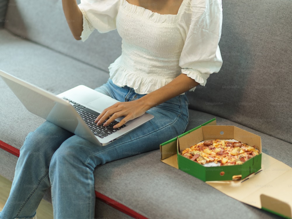 Ausschnittaufnahme einer Frau, die von zu Hause aus mit Laptop und Pizza auf dem Sofa im Wohnzimmer arbeitet