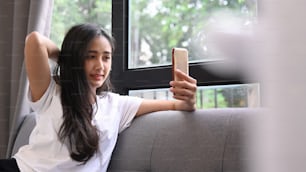 Giovane donna allegra seduta sul divano di casa e con in mano lo smartphone che fa selfie.