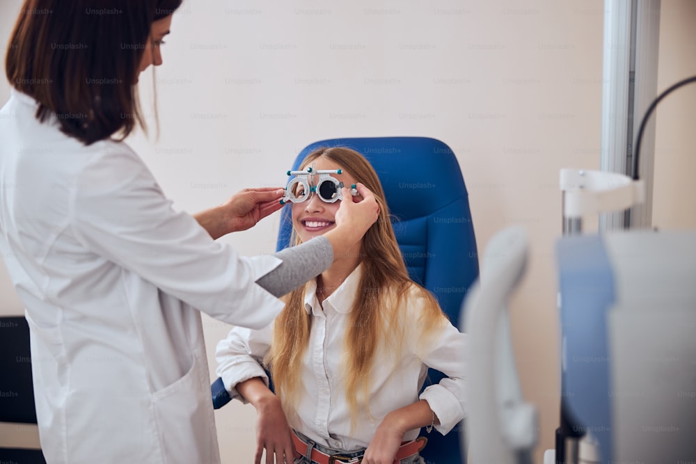 Bonita joven con ropa informal sentada en la silla de la medicina mientras el médico examina los marcos de prueba para comprobar la vista en la clínica de oftalmología moderna