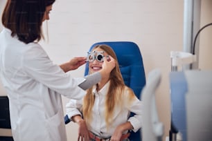 Hübsche junge Dame in Freizeitkleidung sitzt auf einem Medizinstuhl, während der Arzt in der modernen Augenklinik Proberahmen zur Überprüfung der Sehkraft untersucht