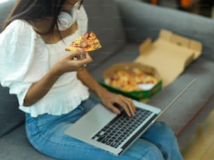 Ausschnittaufnahme eines weiblichen Teenagers, der Pizza isst, während sie den Laptop auf ihrem Schoß benutzt, während sie auf der Couch sitzt