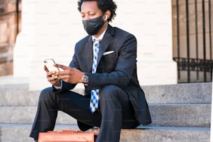 얼굴 마스크를 쓰고 거리에서 야외 계단에 앉아 휴대폰을 사용하는 전문 사업가. 새로운 정상적인 라이프 스타일. 사업 개념입니다.