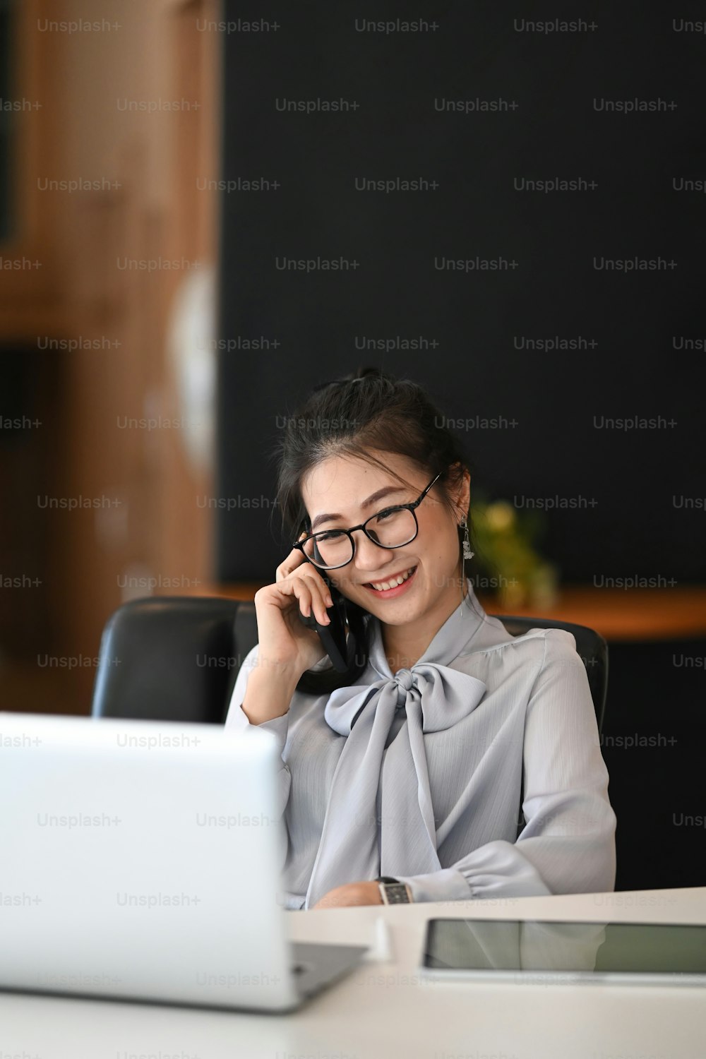 Portrait of businesswoman in eyeglasses working with laptop computer and talking on mobile phone in modern office.