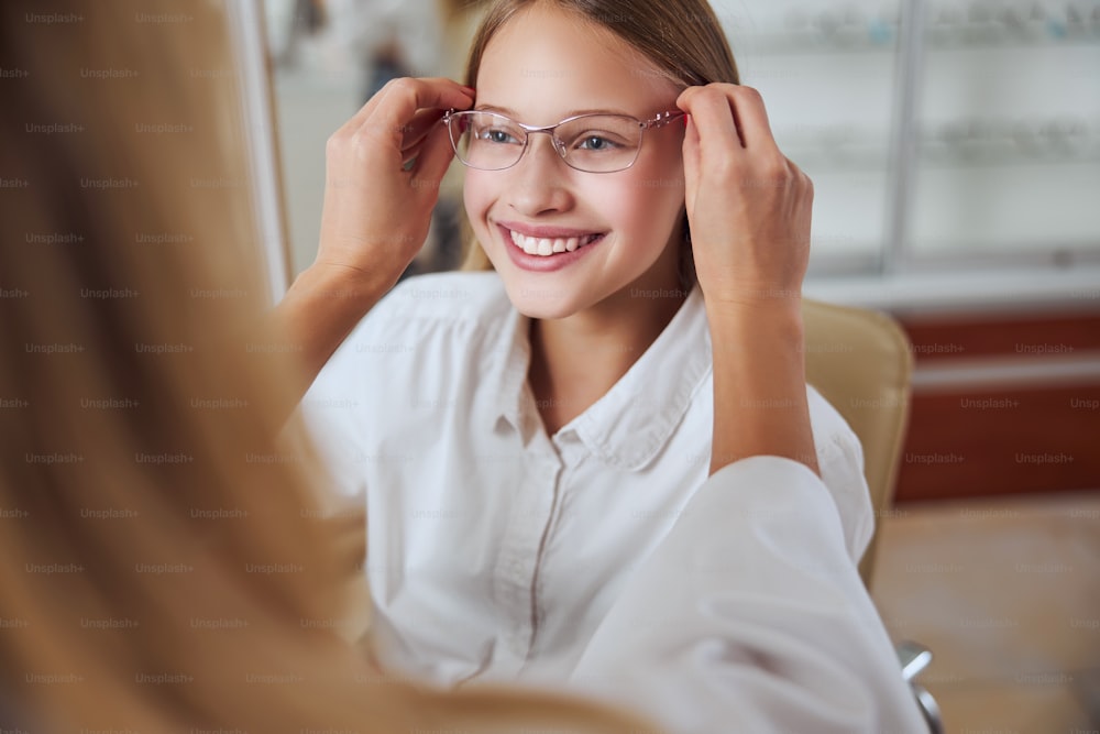 Close up portrait of happy smiling teenager girl in white shirt wearing eyeglass while sitting in the chair in optician clinic