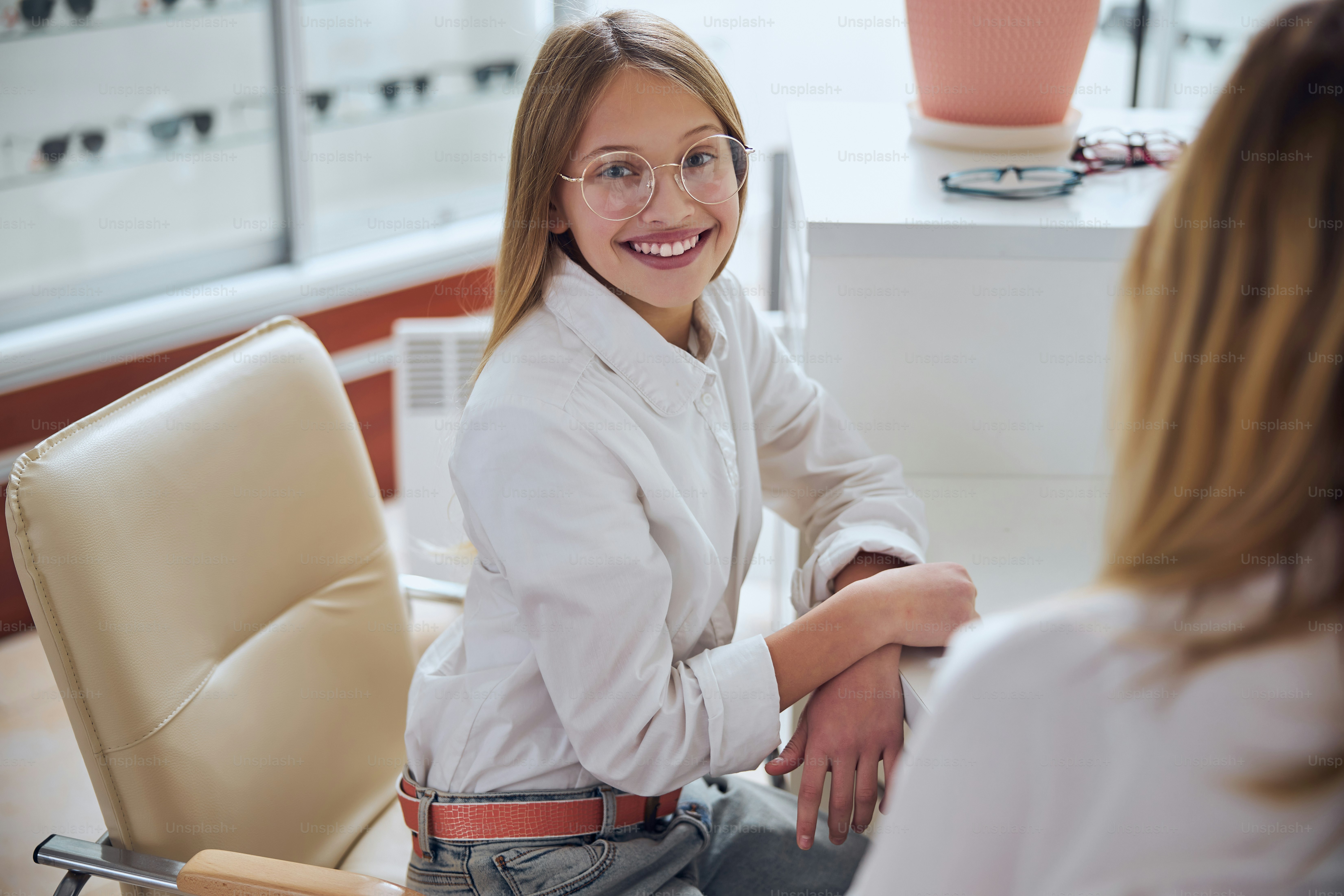 Portrait à la taille d’une jeune femme joyeuse et heureuse en chemise blanche assise à la table tout en regardant l’appareil photo dans un magasin d’optique