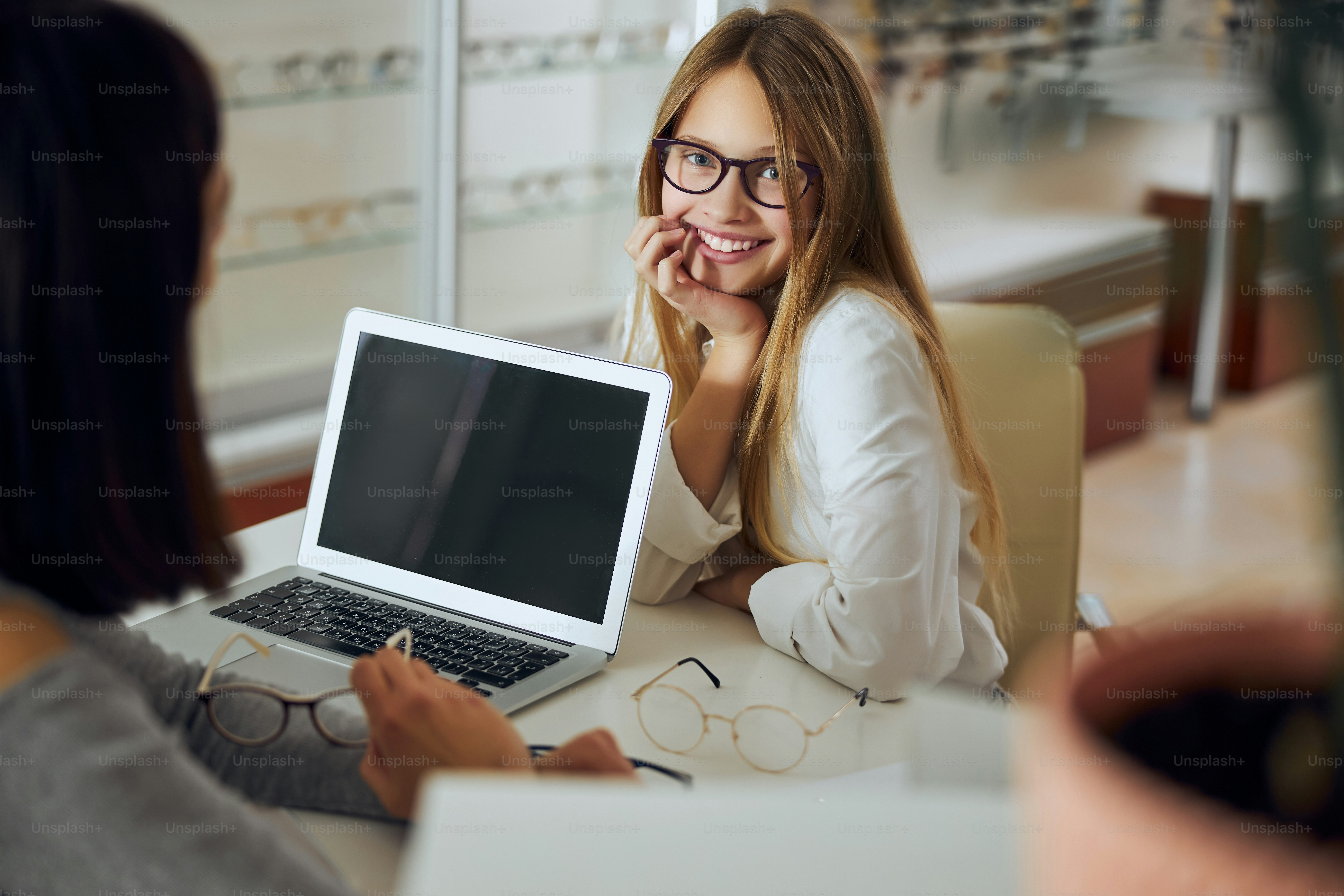 Portrait de taille d’une adolescente élégante assise au bureau et essayant différents verres dans un centre d’opticien tout en parlant avec une vendeuse