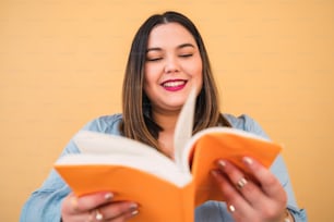 Portrait of young plus size woman enjoying free time and reading a book while standing against yellow background. Lifestyle concept.