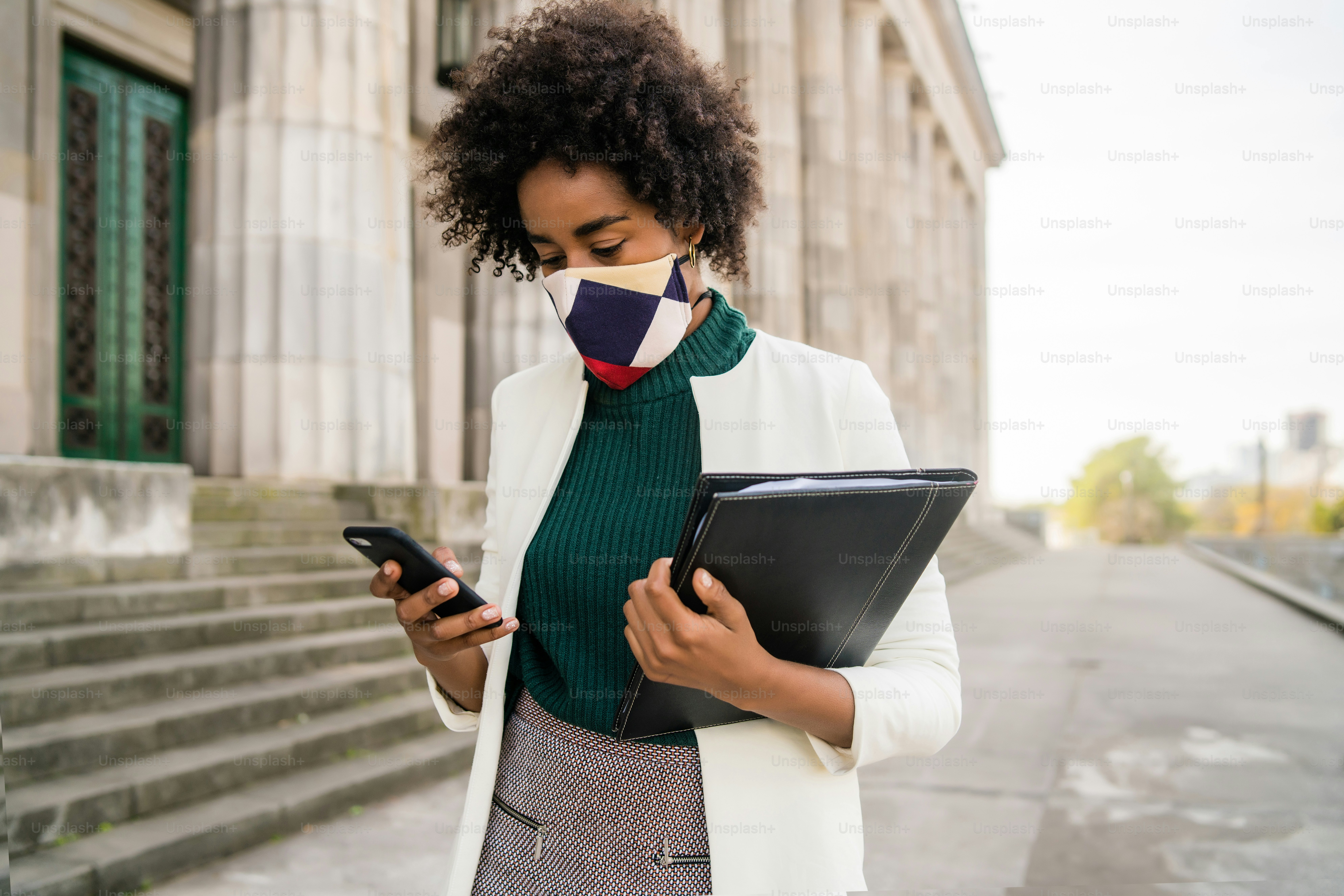 Portrait d’une femme d’affaires afro avec un masque de protection et utilisant son téléphone portable tout en se tenant à l’extérieur dans la rue. Concept d’entreprise et urbain.