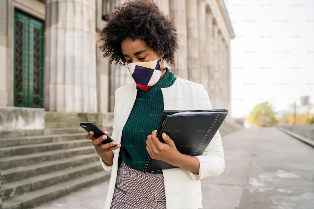 Portrait of afro business woman with protective mask and using her mobile phone while standing outdoors at the street. Business and urban concept.