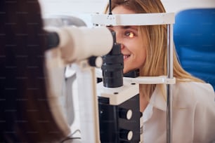 Close up side view portrait of beautiful smiling female looking at slip lamp machine in optometrist cabinet